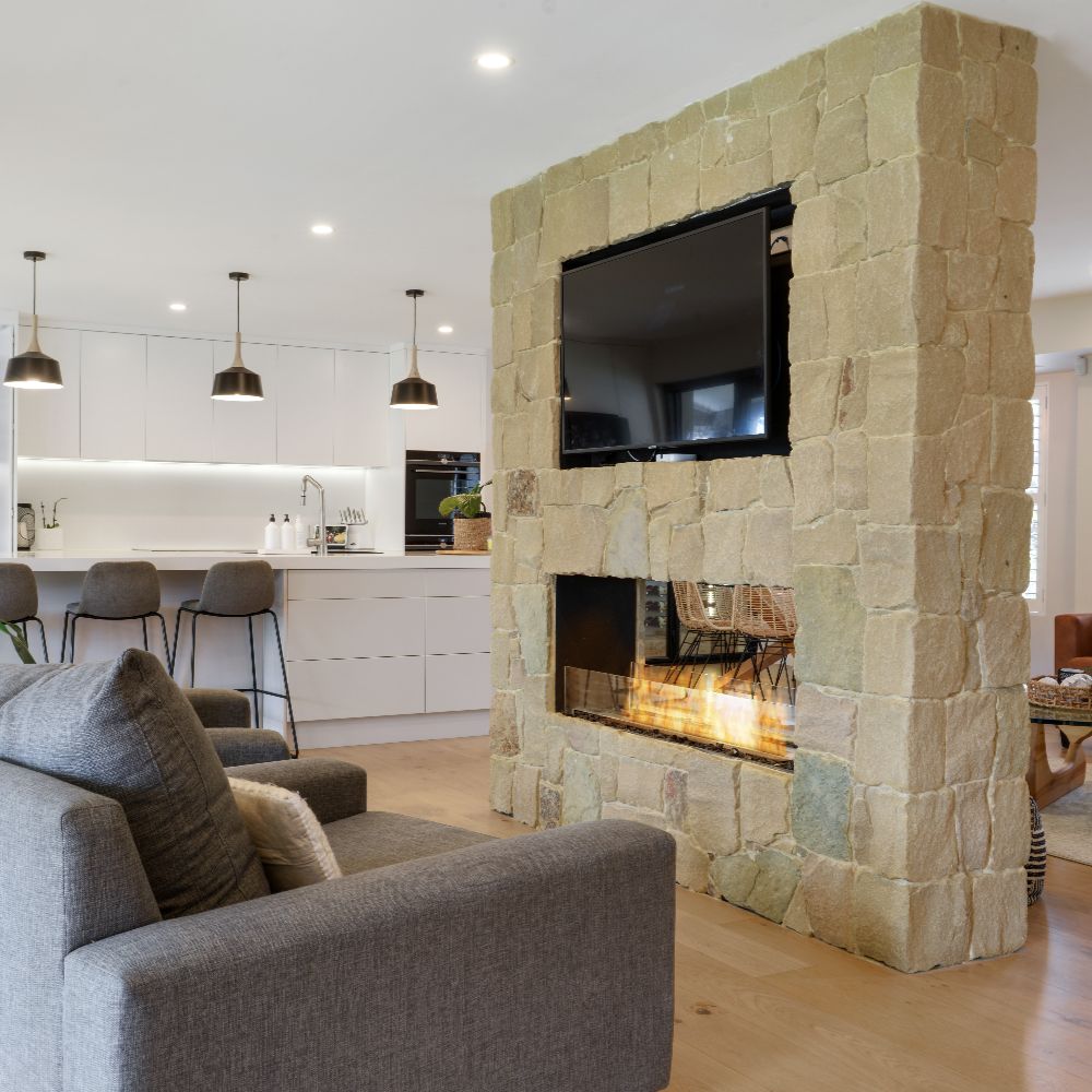Living room with stone fireplace and TV, kitchen in the background
