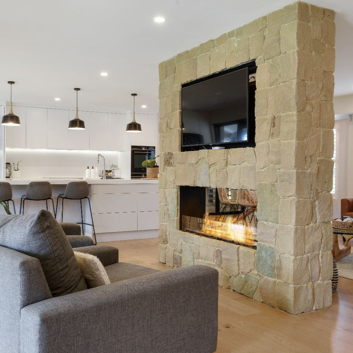 Living room with stone fireplace and TV, kitchen in the background